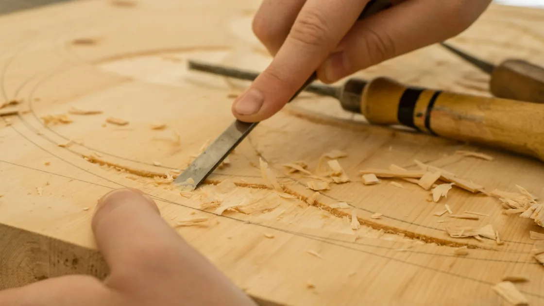 Gubia de filo recto abriendo una línea curva de ornamento sobre una tabla de madera clara, con virutas y otras herramientas alrededor